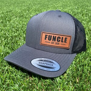 May include: A gray and black baseball cap with a brown leather patch that says "FUNCLE EST. 2024". The cap has a mesh back and a black and silver sticker that says "THE AUTHENTIC YUPOONG SNAPBACK YUPOONG SINCE 1974".
