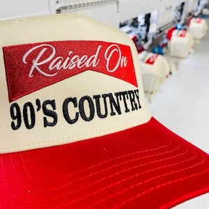May include: A beige and red baseball cap with the text "Raised On 90's Country" embroidered on the front. The cap has a red brim and a beige crown. The text is in white and black.