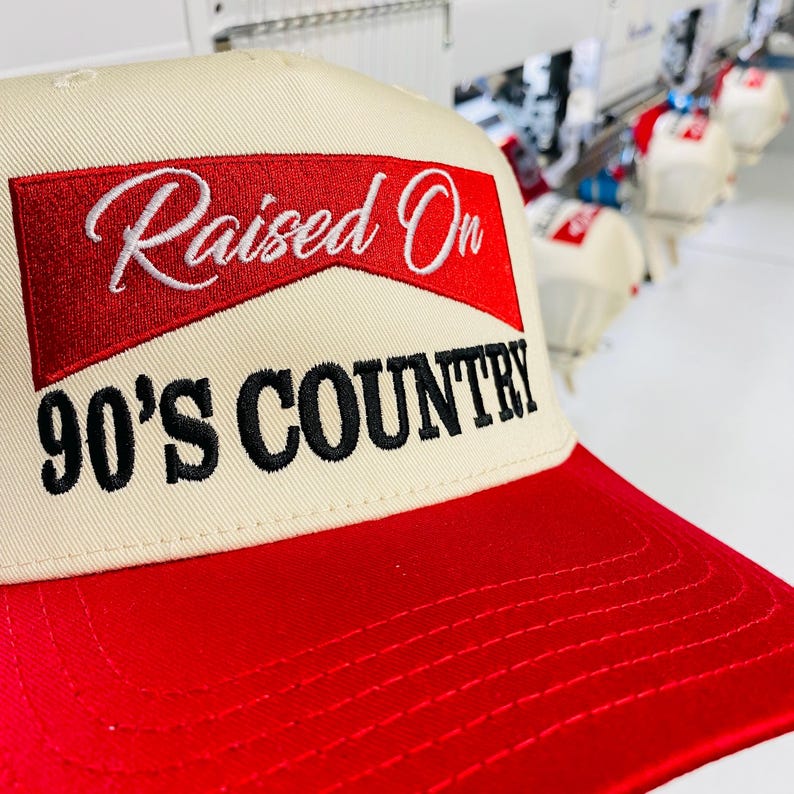 May include: A beige and red baseball cap with the text "Raised On 90's Country" embroidered on the front. The cap has a red brim and a beige crown. The text is in white and black.