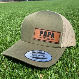 May include: A green and beige baseball cap with a brown leather patch that says "PAPA EST. 2024". The cap has a silver sticker that says "THE AUTHENTIC YUPOONG SNAPBACK YUPOONG SINCE 1974".