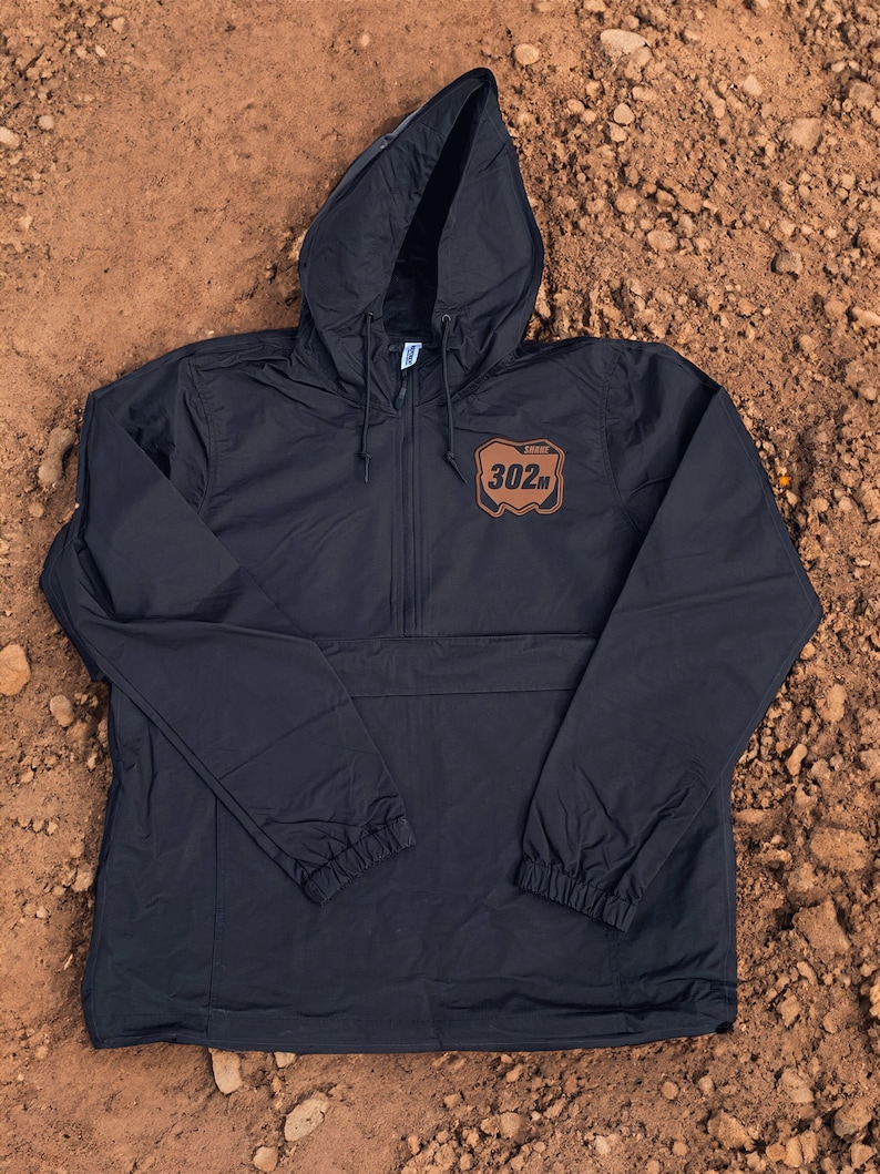Puede incluir: Una chaqueta cortavientos negra tipo pullover con capucha y cordones, mangas largas con pu&ntilde;os el&aacute;sticos y un parche marr&oacute;n en el pecho que muestra el texto 302M y SHANT en letras negras. La chaqueta tiene un cierre de media cremallera frontal.