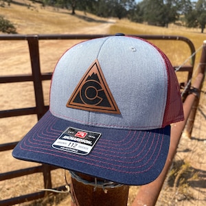 May include: A gray and navy blue trucker hat with a brown leather patch featuring a mountain and a letter C. The hat has a red mesh back and a black label that reads "Richardson 112 Trucker Adjustable".