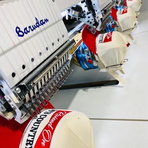 May include: An industrial view of embroidery machines in operation, stitching baseball caps. The caps are cream-colored with red brims and feature the text "Raised On Country". The machines have the name "Barudan" embroidered in blue.
