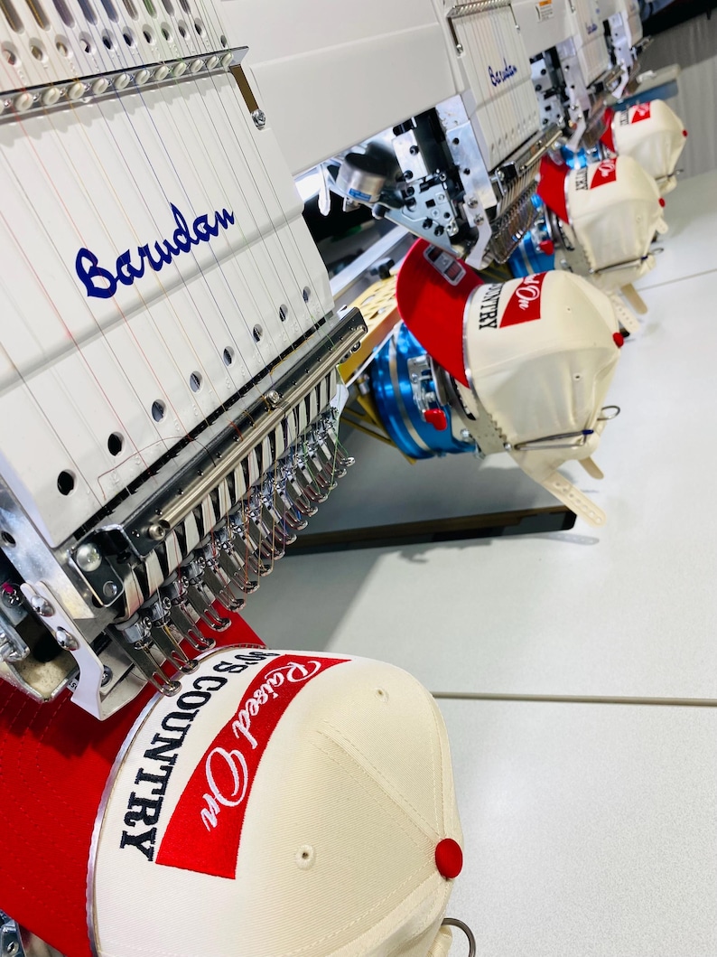 May include: An industrial view of embroidery machines in operation, stitching baseball caps. The caps are cream-colored with red brims and feature the text "Raised On Country". The machines have the name "Barudan" embroidered in blue.