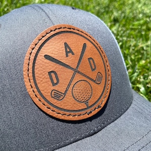 May include: A gray baseball cap with a brown leather patch. The patch features the word "DAD" with crossed golf clubs and a golf ball. The cap has a mesh back.