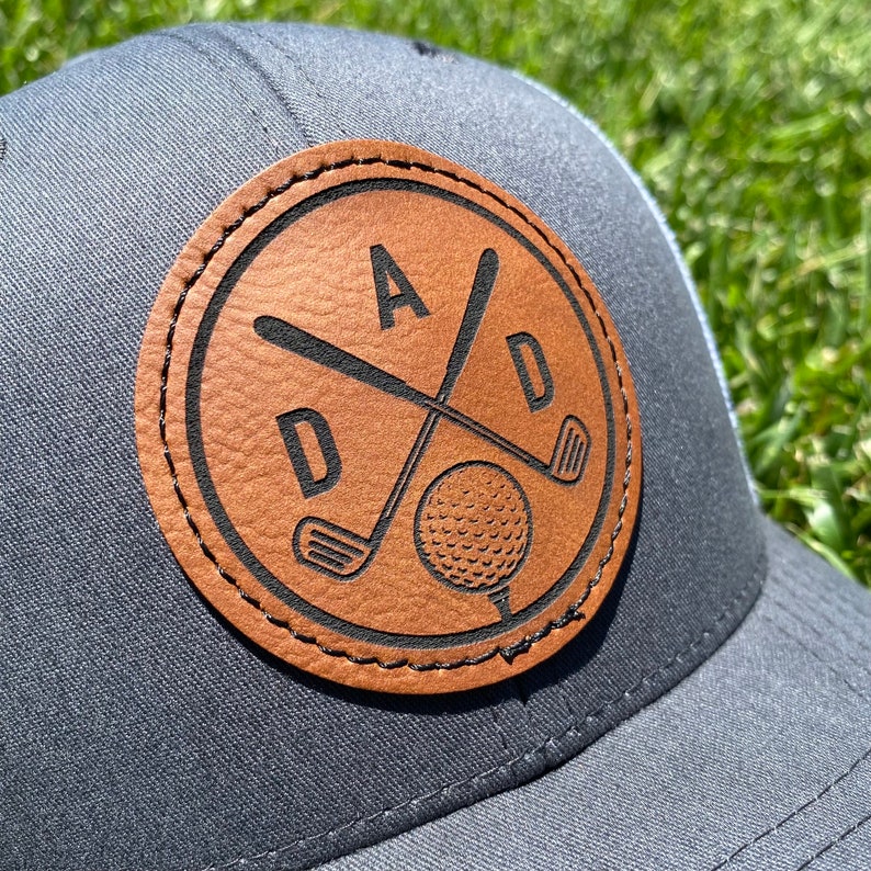 May include: A gray baseball cap with a brown leather patch. The patch features the word "DAD" with crossed golf clubs and a golf ball. The cap has a mesh back.