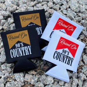 May include: Black and white beverage can coolers with the text "Raised On 90's Country" in a stylized design. The coolers are made of a fabric material and are designed to keep drinks cold. The background is a bed of small gray rocks.