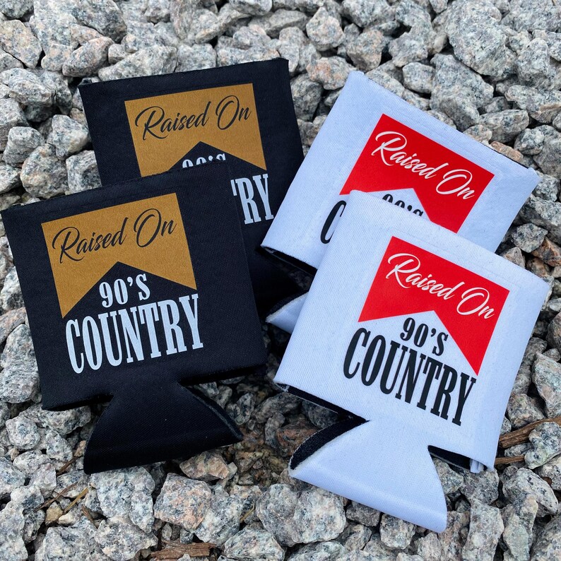 May include: Black and white beverage can coolers with the text "Raised On 90's Country" in a stylized design. The coolers are made of a fabric material and are designed to keep drinks cold. The background is a bed of small gray rocks.