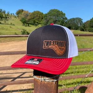 May include: A black and white trucker hat with a red brim. The hat has a brown leather patch with the text "TWO STROKE" and a stylized image of a motorcycle exhaust pipe.