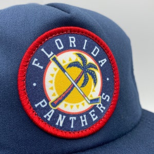 May include: A navy blue trucker hat with a red, white, and yellow embroidered patch that says "Florida Panthers". The patch features a palm tree and two hockey sticks crossed.