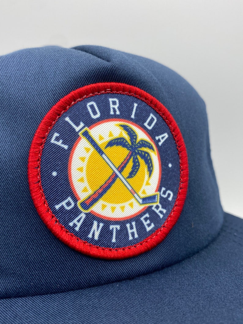 May include: A navy blue trucker hat with a red, white, and yellow embroidered patch that says "Florida Panthers". The patch features a palm tree and two hockey sticks crossed.