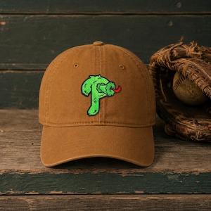 May include: A brown baseball cap featuring an embroidered green letter P with a red tongue sticking out. The cap has a structured crown, a curved brim, and eyelets for ventilation. The design is centered on the front panel of the hat.