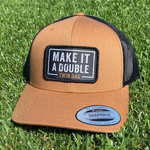 May include: A brown and black trucker hat with the text "MAKE IT A DOUBLE TWIN DAD" on a black and white patch. The hat has a curved brim and a mesh back. The hat is sitting on green grass.