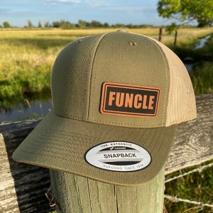 May include: An olive green and tan FUNCLE snapback hat with a leather patch. The patch has the word "FUNCLE" in bold, black letters. The hat has a mesh back and a curved brim. The hat is sitting on a wooden post.