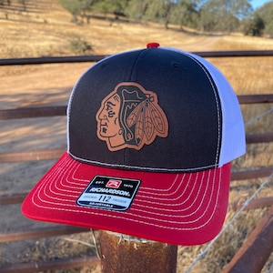 May include: Black and white trucker hat with a red brim. The hat features a brown leather patch with a Native American headdress and the Chicago Blackhawks logo. The hat is labeled "Richardson 112 Trucker Adjustable".