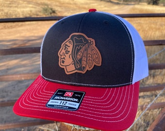 May include: Black and white trucker hat with a red brim. The hat features a brown leather patch with a Native American headdress and the Chicago Blackhawks logo. The hat is labeled "Richardson 112 Trucker Adjustable".