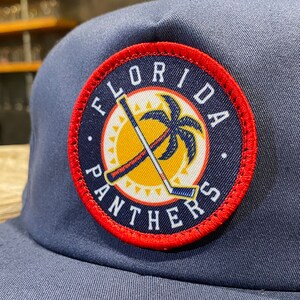 May include: A navy blue baseball cap with a red, white, and yellow embroidered patch that says "Florida Panthers". The patch features a palm tree and two hockey sticks crossed over each other.