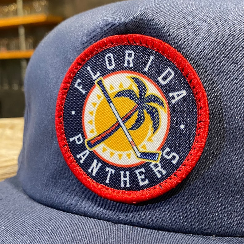 May include: A navy blue baseball cap with a red, white, and yellow embroidered patch that says "Florida Panthers". The patch features a palm tree and two hockey sticks crossed over each other.