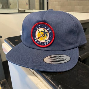May include: A navy blue baseball cap with a red, white, and yellow embroidered patch that says "Florida Panthers". The patch features a palm tree and two crossed hockey sticks. The cap has a black and silver sticker on the back that says "The Authentic Yupoong Snapback Since 1974".