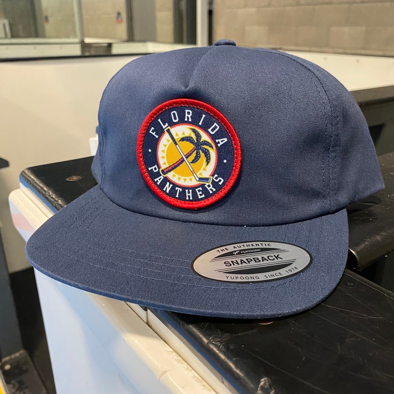 May include: A navy blue baseball cap with a red, white, and yellow embroidered patch that says "Florida Panthers". The patch features a palm tree and two crossed hockey sticks. The cap has a black and silver sticker on the back that says "The Authentic Yupoong Snapback Since 1974".