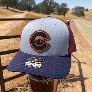 May include: A gray and red trucker hat with a brown leather patch featuring a mountain range and the letter C. The hat is sitting on a rusty metal fence post. The hat has a Richardson 112 adjustable trucker label.