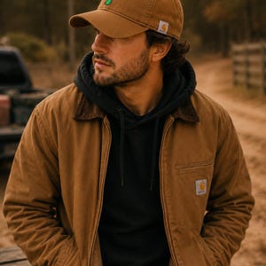 May include: A brown baseball cap with a small green creature graphic, a black hooded sweatshirt, and a brown canvas jacket with a dark brown collar. The cap and jacket feature a small white and orange square logo patch with the text Carhartt.