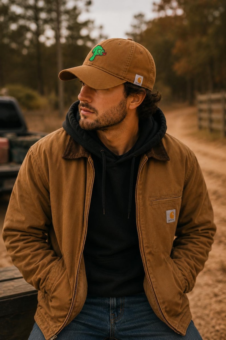 May include: A brown baseball cap with a small green creature graphic, a black hooded sweatshirt, and a brown canvas jacket with a dark brown collar. The cap and jacket feature a small white and orange square logo patch with the text Carhartt.