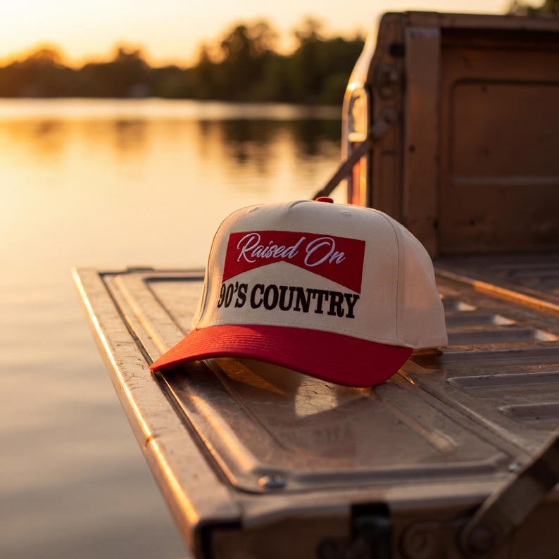 May include: A baseball cap with a white crown and a red brim. The cap features the text "Raised On 90's Country" in a red and white design. The hat is resting on a metal surface.