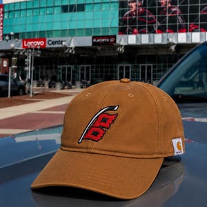 Puede incluir: Gorra de b&eacute;isbol marr&oacute;n con un logotipo rojo y blanco, colocada sobre un coche. La gorra tiene una visera curva y un logotipo de Carhartt. El fondo muestra un edificio con un letrero de Lenovo y jugadores de hockey.