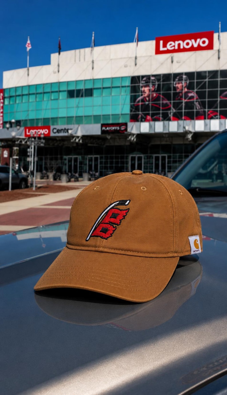 Puede incluir: Gorra de b&eacute;isbol marr&oacute;n con un logotipo rojo y blanco, colocada sobre un coche. La gorra tiene una visera curva y un logotipo de Carhartt. El fondo muestra un edificio con un letrero de Lenovo y jugadores de hockey.