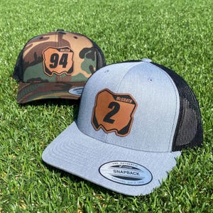 May include: Two black and grey trucker hats with a brown leather patch featuring a number and name. The hat on the left has the number 94 and the name "Roczen". The hat on the right has the number 2 and the name "McGrath".