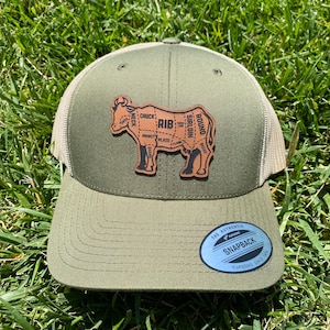 May include: A green and beige trucker hat with a brown leather patch depicting a cow with different cuts of meat labeled. The patch reads "SIRLOIN", "ROUND", "SHANK", "CHUCK", "RIB", "BRISKET", "PLATE", "FLANK", and "NECK". The hat has a white label that reads "THE AUTHENTIC YUPOONG SNAPBACK YUPOONG SINCE 1974".