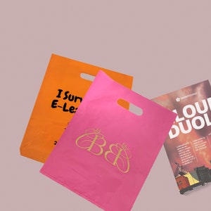 May include: Two pink and orange reusable shopping bags with a gold logo and the text "I Sur E-Le" on the orange bag. A magazine with the text "LOU DUOL" is also visible.