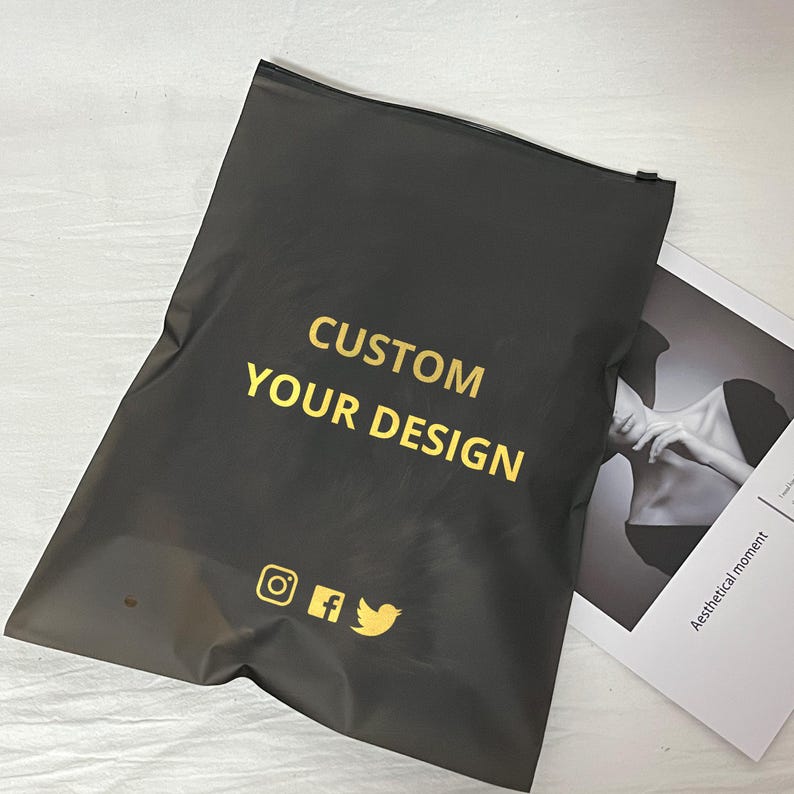 Peut inclure: Un sac d'exp&eacute;dition noir avec le texte "CUSTOM YOUR DESIGN" en or. Le sac pr&eacute;sente &eacute;galement des ic&ocirc;nes de m&eacute;dias sociaux dor&eacute;es pour Instagram, Facebook et Twitter. Un magazine avec une photo en noir et blanc est partiellement visible.