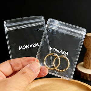 May include: Two gold-colored hoop earrings with a small, clear gemstone are displayed in a clear plastic bag. The bag has a zip closure and the text "MONA2M" printed on it. The earrings have a textured design.
