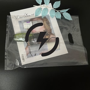May include: A clear plastic bag with a handle, containing a magazine with a woman on the cover and several bottles of beauty products. The magazine has the word "Vulture" written on the cover. The bag has a black lightning bolt symbol on it.