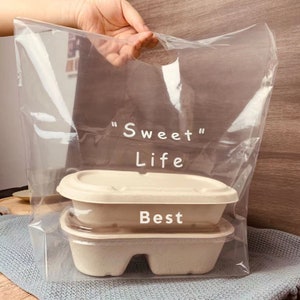 May include: A clear plastic bag with a handle, printed with the text "Sweet Life" and "Best". The bag contains two stacked brown cardboard containers.