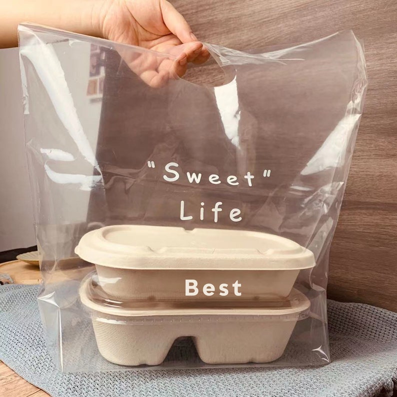 May include: A clear plastic bag with a handle, printed with the text "Sweet Life" and "Best". The bag contains two stacked brown cardboard containers.