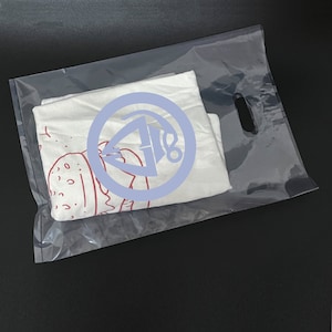 May include: A white t-shirt with a red and white graphic of a burger and a blue circle with a red graphic inside. The t-shirt is in a clear plastic bag with a handle.