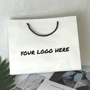 Puede incluir: Bolsa de papel blanca con asas de cuerda negra y el texto "YOUR LOGO HERE" impreso en negro.