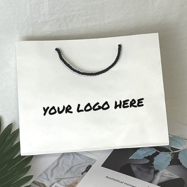 Puede incluir: Bolsa de papel blanca con asas de cuerda negra y el texto "YOUR LOGO HERE" impreso en negro.
