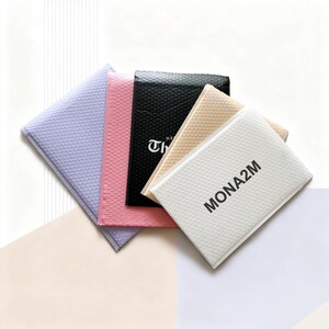 Puede incluir: Una colecci&oacute;n de sobres de burbujas de colores, en tonos lavanda, rosa, negro, beige y blanco. El sobre blanco muestra el texto "MONA2M" en negro, mientras que el sobre negro tiene el texto "THE" en blanco.