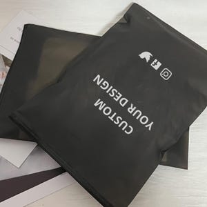 Peut inclure: Deux sacs d'exp&eacute;dition noirs en poly&eacute;thyl&egrave;ne avec le texte blanc "CUSTOM YOUR DESIGN". Les sacs comportent &eacute;galement des ic&ocirc;nes de m&eacute;dias sociaux. Les sacs sont superpos&eacute;s.