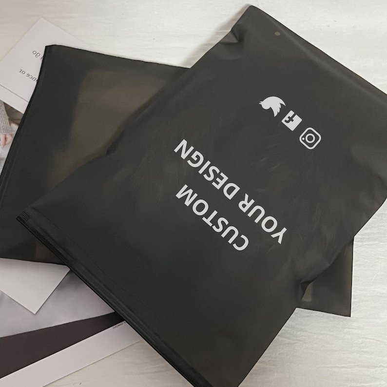 Peut inclure: Deux sacs d'exp&eacute;dition noirs en poly&eacute;thyl&egrave;ne avec le texte blanc "CUSTOM YOUR DESIGN". Les sacs comportent &eacute;galement des ic&ocirc;nes de m&eacute;dias sociaux. Les sacs sont superpos&eacute;s.