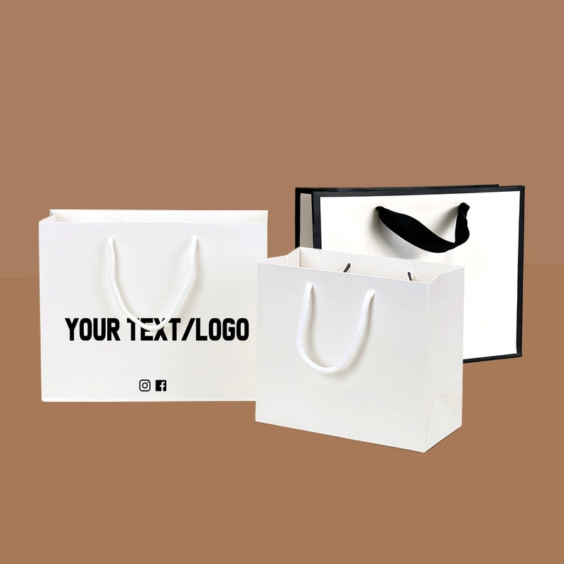 Puede incluir: Tres bolsas de papel blancas con asas de cuerda. Dos bolsas tienen asas de cuerda blancas y una bolsa tiene un asa de cuerda negra. La bolsa de la izquierda tiene el texto "YOUR TEXT/LOGO" impreso en la parte delantera. La bolsa de la derecha tiene un borde negro alrededor de la parte superior e inferior.