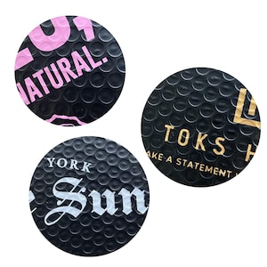 Puede incluir: Tres pegatinas circulares negras con una superficie texturizada. Una pegatina tiene la palabra "NATURAL" en rosa. Otra tiene "TOKS" y "MAKE A STATEMENT" en dorado. La tercera tiene "YORK Sun" en blanco.