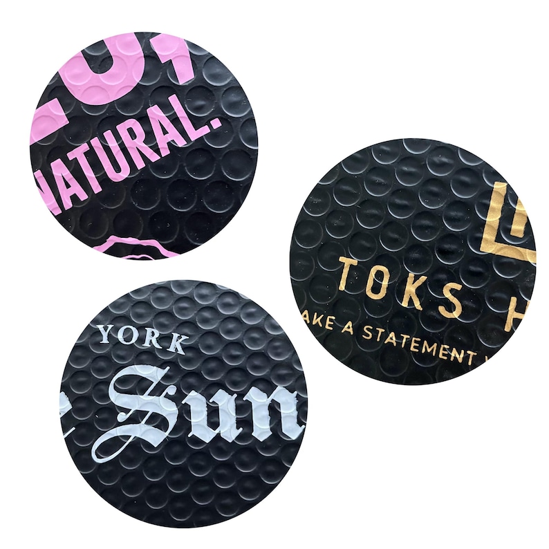 Puede incluir: Tres pegatinas circulares negras con una superficie texturizada. Una pegatina tiene la palabra "NATURAL" en rosa. Otra tiene "TOKS" y "MAKE A STATEMENT" en dorado. La tercera tiene "YORK Sun" en blanco.