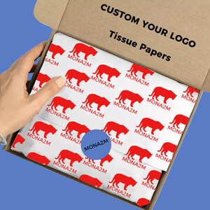 May include: A white box with a red tiger pattern and the text "CUSTOM YOUR LOGO Tissue Papers" on the top. The box is open and a hand is reaching in to grab a sheet of tissue paper. The tissue paper has a blue circle with the text "MONA2M" in white.