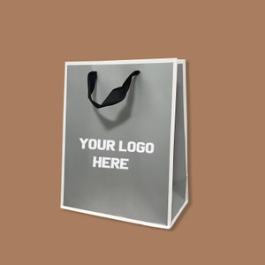 Puede incluir: Una bolsa de papel de color gris con un borde blanco y asas negras. La bolsa tiene el texto "YOUR LOGO HERE" impreso en la parte delantera.
