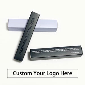 Puede incluir: Cajas de brillo labial negras con las palabras "HANDMADE LIPGLOSS" impresas en el lateral. Las cajas son rectangulares y tienen un acabado mate. La caja inferior tiene los ingredientes enumerados. El texto "Custom Your Logo Here" está en la parte inferior.